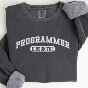 Op de afbeelding: Een donkergrijs sweatshirt met de tekst "PROGRAMMER" in een witte boog boven de tekst "DIBS ON THE" in een witte ovaal. Het sweatshirt heeft een fleecevoering.