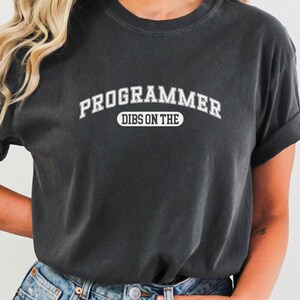 May include: A dark grey t-shirt with white text that reads "PROGRAMMER DIBS ON THE" in an arched font.