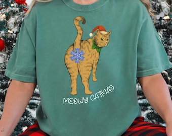 Puede incluir: Camiseta verde con un gato de dibujos animados con un gorro de Papá Noel y un copo de nieve en la parte trasera. El texto "MEOWY CATMAS" está impreso en blanco. La camiseta se combina con un pantalón de pijama a cuadros rojos y negros.
