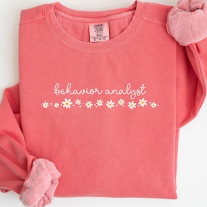 May include: A watermelon-coloured sweatshirt with the text "behaviour analyst" in white script with a row of white daisies below.