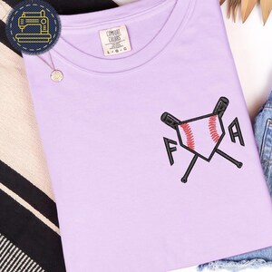 Op de afbeelding: Een lichtpaars t-shirt met een geborduurd honkbalthema. Het ontwerp toont een honkbal, gekruiste knuppels en de letters "F" en "A". Het shirt is opgevouwen en op een wit oppervlak geplaatst.