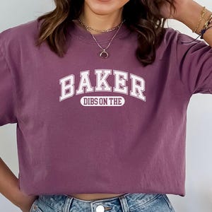 May include: A mauve colored t-shirt with white text that reads "BAKER DIBS ON THE".