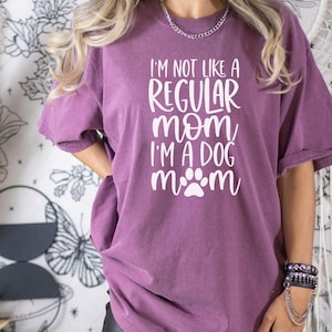 May include: A purple t-shirt with the text "I'm not like a regular mom, I'm a dog mom" in white lettering with a paw print.