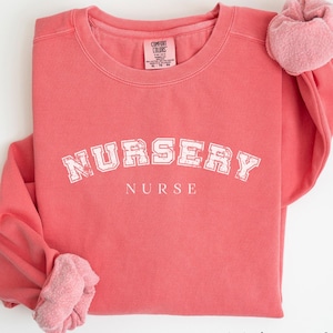 May include: Watermelon colored sweatshirt with the text "Nursery Nurse" in a white collegiate font.