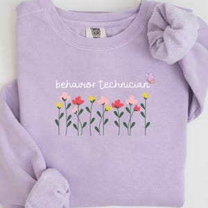 Könnte beinhalten: Ein helllila Sweatshirt mit dem weißen Schriftzug "behavior technician". Darunter eine Reihe bunter Blumen und ein kleiner Schmetterling. Das Wort "Orchid" steht in Schreibschrift unten rechts.