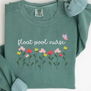 Peut inclure: Sweat-shirt vert clair avec le texte "float pool nurse" et une rangée de fleurs colorées et un papillon.