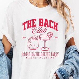 May include: White t-shirt with a red graphic that reads "THE BACH CLUB" above two cocktail illustrations. Below the graphic, the text reads "EMMA'S BACHELORETTE PARTY MIAMI, FLORIDA". The shirt is worn with a denim jacket.