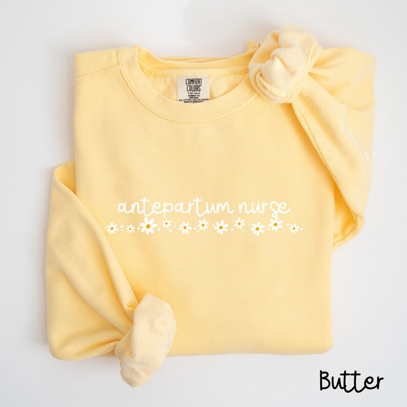 Pode incluir: Uma camisa de moletom amarela com o texto "antepartum nurse" e uma fileira de margaridas brancas impressas na frente.