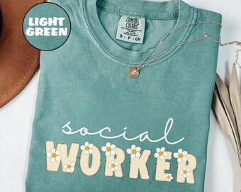 Può includere: Una maglietta verde chiaro con il testo "social worker" in scrittura bianca. La parola "worker" è in grandi lettere gialle con accenti di margherita bianca.