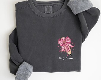 Puede incluir: Una sudadera gris oscuro con un lápiz rosa con un lazo en él y el texto "Mrs. Brown" impreso en la parte delantera. La sudadera tiene un forro de vellón en el interior de las mangas.