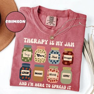 Puede incluir: Una camiseta rosa con el texto "Therapy is my jam" y una fila de tarros con palabras como "Tiny", "Care", "Heal", "Snuggle", "Grow", "Bright", "Brave" y "Giggle". El texto "And I'm here to spread it" está debajo de los tarros.