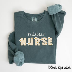 Könnte beinhalten: Ein blaugrüner Pullover mit dem Text "nicu NURSE" in Weiß mit gelben Gänseblümchen-Akzenten.