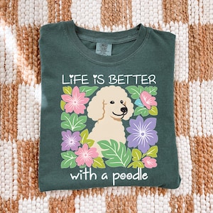 May include: Dark green t-shirt with the text "LIFE IS BETTER with a poodle." The shirt features a cream-colored poodle illustration surrounded by pink, purple, and blue flowers and green leaves. The shirt is made of a soft material.