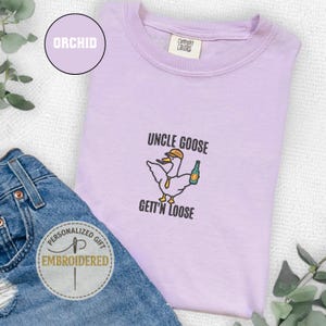 以下が含まれることがあります： 「UNCLE GOOSE GETT'N LOOSE」の刺繍と、帽子とボトルを持った漫画のガチョウが特徴の薄紫色のTシャツ。青いジーンズと「Personalized Gift Embroidered」のラベルと組み合わせています。
