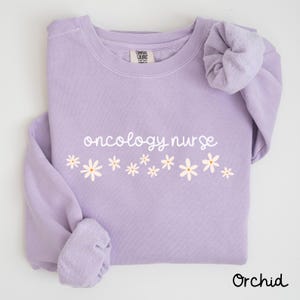 May include: A lavender sweatshirt with the text "oncology nurse" in white script and a row of white daisies. The sweatshirt has a crew neck and long sleeves.