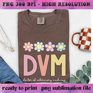 May include: A brown t-shirt with the letters "DVM" in pastel colors and floral accents. The text "doctor of veterinary medicine" is below the letters. A mug with a brown and white checkered pattern is on the right.