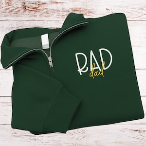 May include: A dark green quarter-zip sweatshirt with the words "RAD dad" embroidered in white and yellow. The sweatshirt is folded on a white wooden surface. The zipper is silver.