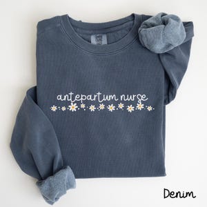 Pode incluir: Uma camisa de moletom azul escura com o texto "antepartum nurse" em letras brancas e uma fileira de margaridas brancas com centros amarelos.