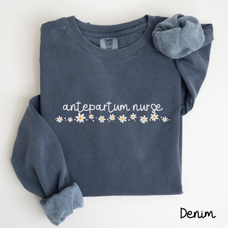 Pode incluir: Uma camisa de moletom azul escura com o texto "antepartum nurse" em letras brancas e uma fileira de margaridas brancas com centros amarelos.