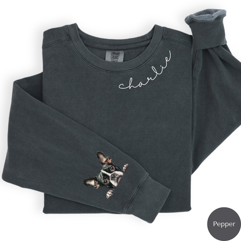 Pode incluir: Uma camisa de moletom cinza escuro com o nome "charlie" bordado na frente e um cachorro preto e branco espiando da manga. A camisa de moletom &eacute; feita de um tecido macio e confort&aacute;vel.