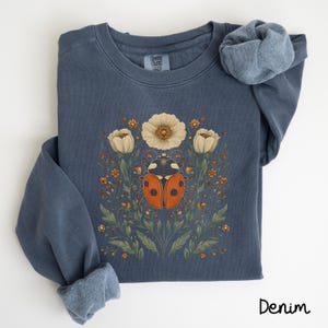 May include: A dark blue sweatshirt with a graphic of a ladybug surrounded by white flowers and green leaves. The text "COMFORT COLORS" is visible on the tag.