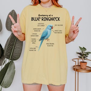 May include: Yellow t-shirt with the text "Anatomy of a Blue Ringneck" and an illustration of a blue bird with labeled anatomical features. The bird has a red beak and is labeled with humorous descriptions like "Spicy Chili Beak".