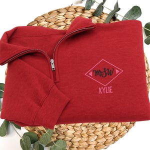 May include: A red quarter-zip sweatshirt with a silver zipper. The sweatshirt features a pink and black embroidered diamond design with the initials "MSW" and the name "KYLIE" below. The sweatshirt is displayed on a woven basket.