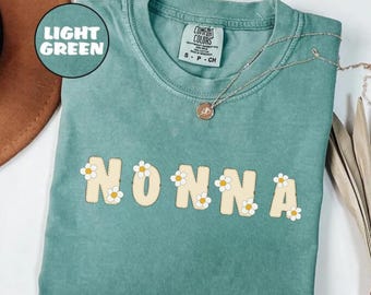 May include: A light green t-shirt with the word "NONNA" in yellow letters with white daisy accents. The shirt is made of a soft, comfortable fabric.