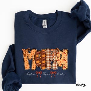 Peut inclure: Sweat-shirt bleu marine avec le mot "MAMA" dans un design sur le thème de l'automne. Les lettres présentent des citrouilles, des feuilles et des motifs à carreaux. Les noms Sophia, Ryan et Emily sont en dessous.
