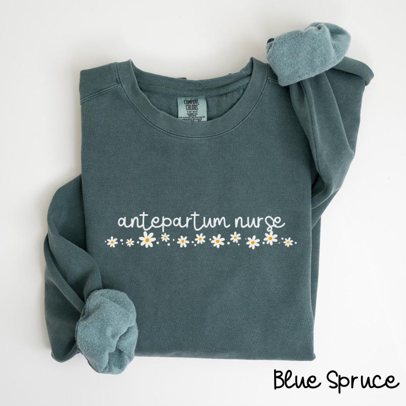 Pode incluir: Uma camisola azul-turquesa com o texto "antepartum nurse" e uma fila de margaridas brancas impressas na frente. A camisola est&aacute; dobrada e as mangas est&atilde;o dobradas.