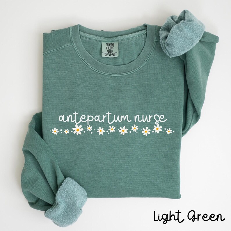 Pode incluir: Camisola verde claro com o texto "antepartum nurse" em branco e uma fila de margaridas brancas abaixo.