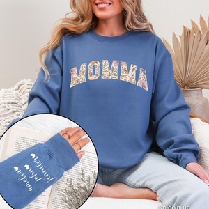 May include: Indigo blue sweatshirt with the word "MOMMA" in a floral design across the chest. The left sleeve has the names "Anelia", "John", and "Jennifer" printed in white with heart symbols. The sweatshirt is a comfortable, casual style.