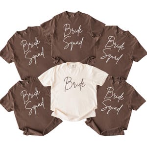 Könnte beinhalten: Sechs T-Shirts, fünf braune mit weißem Schriftzug "Bride Squad" und ein cremefarbenes T-Shirt mit weißem Schriftzug "Bride". Die Shirts sind kreisförmig angeordnet.