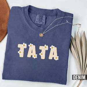 May include: A navy blue t-shirt with the word "TATA" in white letters with daisy accents. The shirt is made of a soft, comfortable material.