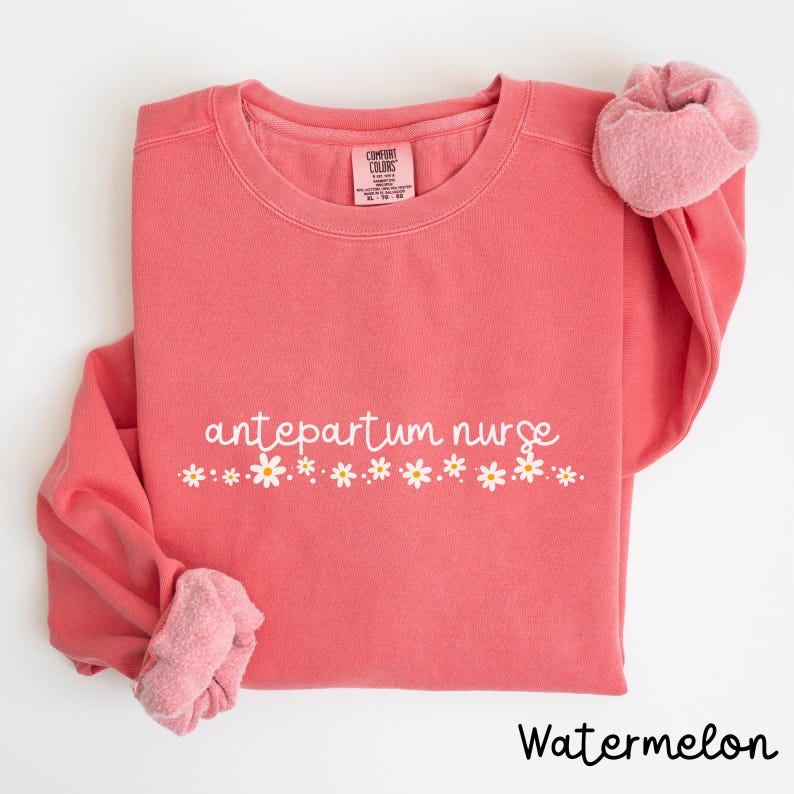 Pode incluir: Uma camisola cor de melancia com o texto "antepartum nurse" em letras brancas com uma fila de margaridas brancas abaixo.