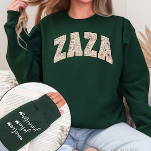 May include: Forest green sweatshirt with the word "ZAZA" in a floral design across the chest. The left sleeve has the names "Amelia", "John", and "Jennifer" in white script with heart symbols. The sweatshirt is a crew neck style.