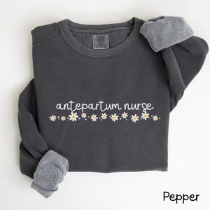 Pode incluir: Uma camisola cinzenta escura com o texto "antepartum nurse" em letras brancas e uma fila de margaridas brancas com centros amarelos.