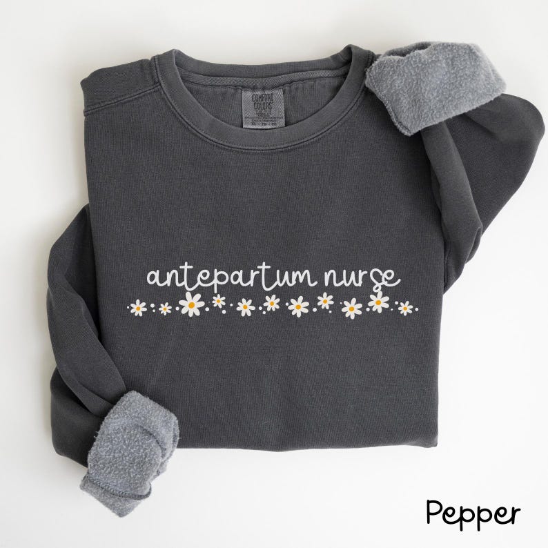 Pode incluir: Uma camisola cinzenta escura com o texto "antepartum nurse" em letras brancas e uma fila de margaridas brancas com centros amarelos.