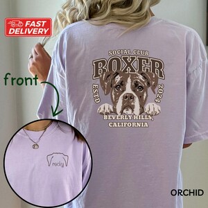May include: A light purple t-shirt with a graphic of a boxer dog and the text "Social Club Boxer EstD 2024 Beverly Hills, California". The front of the shirt has a small embroidered dog paw print with the text "rocky".
