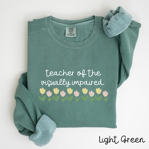 May include: Light green sweatshirt with the text "teacher of the visually impaired" and a row of yellow and pink flowers.