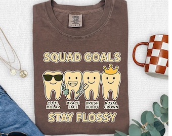 Op de afbeelding: Een bruin T-shirt met de tekst "SQUAD GOALS" en "STAY FLOSSY". Het ontwerp toont vier cartoon tanden, elk met een ander accessoire en gelabeld "COOL MOLAR", "BRACE FACE", "BRUSH BUDDY" en "ROYAL CROWN".