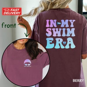 May include: A maroon t-shirt with the text "IN MY SWIM ERA" in a rainbow gradient on the back. The front of the shirt has a small graphic of a swimmer's cap and goggles with the text "IN MY SWIM ERA" underneath.
