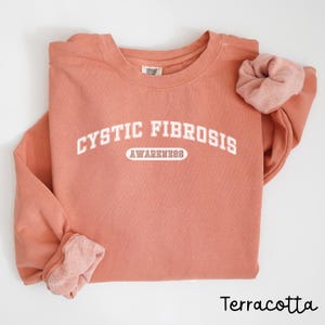 Puede incluir: Sudadera de color terracota con el texto "CYSTIC FIBROSIS AWARENESS" en letras blancas. Una scrunchie a juego descansa sobre la manga. La palabra "Terracota" está escrita en una fuente de guion en la parte inferior.