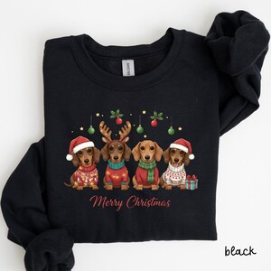 May include: Black sweatshirt with a Christmas design. It features four dachshunds wearing festive sweaters, hats, and reindeer antlers. The words "Merry Christmas" are written in red. The design includes green and red baubles and gold stars, creating a holiday theme.