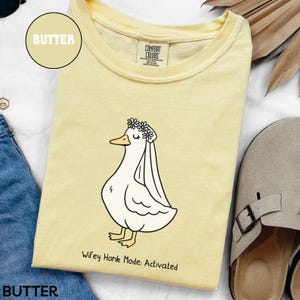 May include: Butter-colored t-shirt with a cartoon duck wearing a floral crown and veil. The text "Wifey Honk Mode: Activated" is printed below the duck. The shirt is made of a soft material.