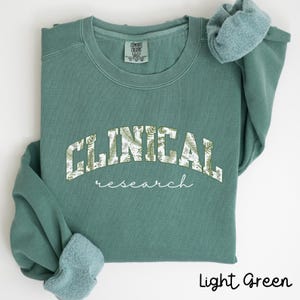 May include: Light green sweatshirt with a white floral design that reads "CLINICAL research" in a vintage collegiate style.