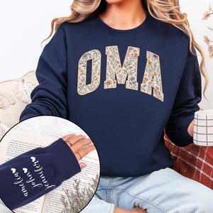 Op de afbeelding: Marineblauw sweatshirt met het woord "OMA" in een bloemenmotief. De sweatshirt heeft een ronde hals en lange mouwen. De manchet van de mouw heeft de namen "Amelia", "John" en "Jennifer" erop.