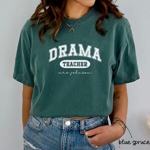 Könnte beinhalten: Ein blaugrünes T-Shirt mit dem Text "Drama Teacher" in Weiß mit einem weißen Oval um das Wort "Teacher" und dem Text "mrs. johnson" in Weiß darunter.