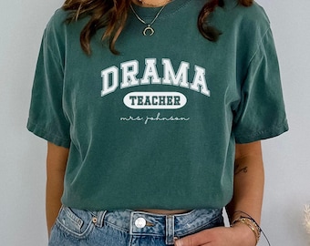 May include: A teal blue t-shirt with the text "Drama Teacher" in white with a white oval around the word "Teacher" and the text "mrs. johnson" in white below.