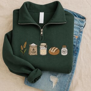 May include: A dark green quarter-zip sweatshirt featuring an embroidered design of wheat stalks, a bag labelled FLOUR, a glass jar, a loaf of bread, and a small jar labelled SALT. The sweatshirt is layered over a pair of blue denim jeans.